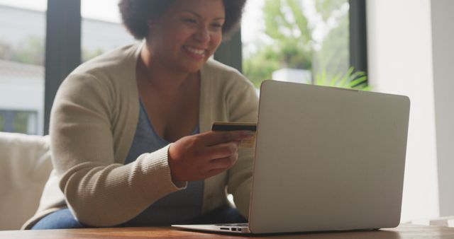 Image of happy plus size african american woman holding credit card and using laptop. lifestyle, leisure, spending free time at home with technology.