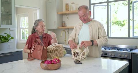 Senior asian couple unpacking reusable mesh grocery bags on bright marble kitchen island