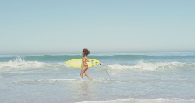 This image shows a young girl running on the beach while holding a yellow surfboard. The sky is clear and the ocean waves are gently crashing. Ideal for summer vacation themes, outdoor activities, surfing promotions, and child-focused advertising.