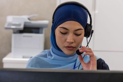 Woman wearing blue headscarf speaking on headset at office computer customer support