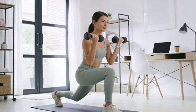 Asian woman lunging with dumbbells in home office wearing sage activewear for fitness