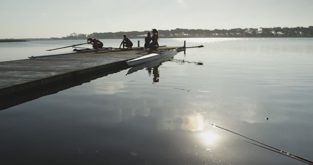 Rowers are depicted sitting and unwinding on a wooden dock as the morning sun rises over a serene lake. This scene, capturing the calm and determination of early morning preparation, could be ideal for representing themes of teamwork, outdoor sports, tranquility, and the beauty of nature. Perfect for websites, blogs, or publications focusing on outdoor activities, sports, fitness, and mindfulness.
