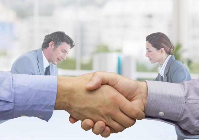 Two business executives in formal attire shaking hands, symbolizing agreement or successful partnership, while two colleagues sit around a table in a meeting. Ideal for concepts of business negotiation, teamwork, corporate collaboration, and agreement.
