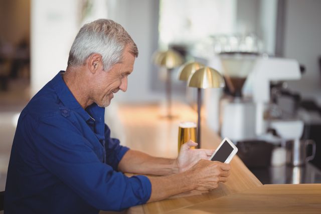 Mature man sitting at a restaurant counter using a digital tablet. He is smiling and appears relaxed, enjoying his time. This image can be used for promoting technology use among seniors, modern lifestyle, leisure activities, or restaurant ambiance.