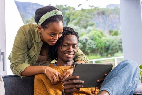 African american couple sharing tablet on sunlit balcony sofa, relaxing and smiling together