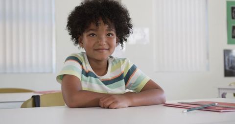 African American boy sitting at classroom desk smiling with red notebook and pencils