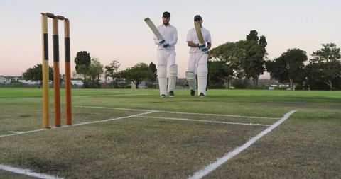 Two diverse batsmen walking onto cricket pitch holding bats near stumps at sunset