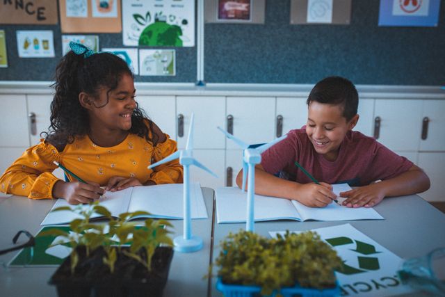 Two diverse schoolchildren are sitting at desks in a classroom, smiling and engaging in an ecology lesson. They are surrounded by educational materials, including wind turbine models and plants, emphasizing sustainability and renewable energy. This image is ideal for use in educational content, environmental campaigns, and materials promoting diversity and inclusion in education.