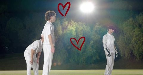 Night cricket players standing under floodlight wearing white kits with red heart overlays