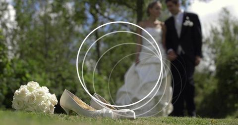 Bridal shoes and white rose bouquet on grass with blurred bride and groom walking
