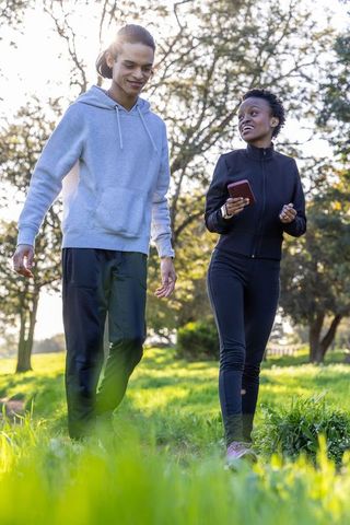 Diverse friends walking and talking in sunlit park wearing athletic wear holding red phone