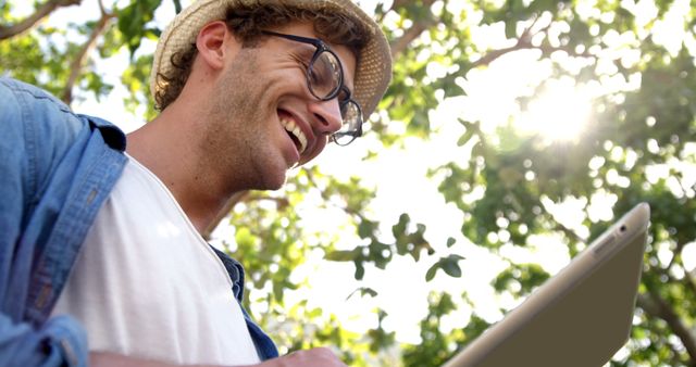 A young Caucasian man smiles as he uses a tablet outdoors, with copy space. His casual attire and the natural setting suggest he might be enjoying leisure time or working remotely in a relaxed environment.
