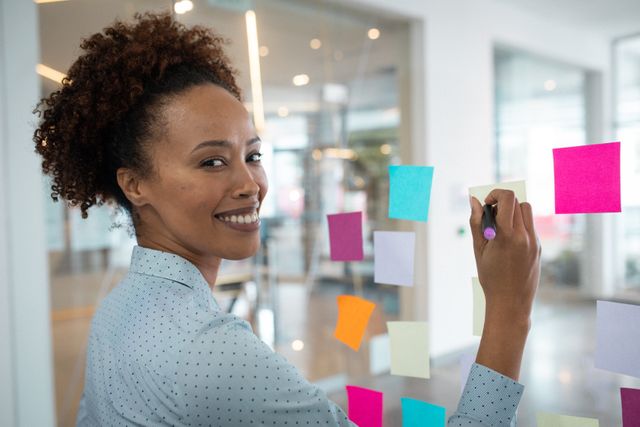 This image depicts a smiling businesswoman writing colorful notes on a glass wall in a modern office. Ideal for illustrating concepts related to brainstorming, planning, teamwork, and creative business environments. Suitable for use in articles, presentations, and marketing materials focusing on professional development, innovation, and collaborative workspaces.