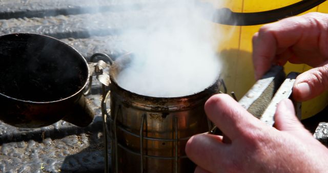 Hands shown lighting a traditional beekeeper's smoker tool with visible smoke and beekeeper equipment. Could be used for agricultural, sustainability, or beekeeper-themed content. Ideal for articles or educational materials on beekeeping practices and tools.