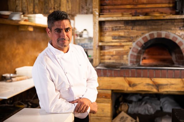 Caucasian male chef standing in a restaurant kitchen, looking at the camera. He is wearing a professional chef's uniform. The background features a rustic wood-fired oven, adding to the culinary atmosphere. Ideal for use in articles or advertisements related to cooking, restaurants, professional chefs, culinary arts, and hospitality industry.