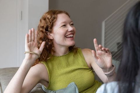 Redhead woman laughing and chatting on sofa wearing green textured top and bracelets