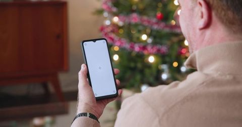 Mature man holding blank-screen smartphone by christmas tree for holiday app mockup