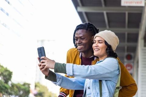 Female and african american male taking selfie at transit shelter, smiling and laughing