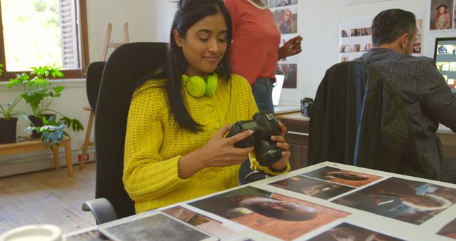Young woman in yellow sweater reviewing photographs in a modern office with camera and headphones, collaborating with colleagues. Ideal for use in marketing materials, creative agency promotions, or technology-focused work environments showing teamwork and project collaboration.