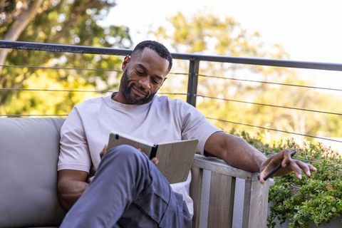 Mature african american man relaxing on balcony reading tablet while holding smartphone
