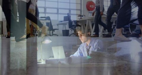 Focused professional reviewing rolled blueprints at laptop in busy open-plan office