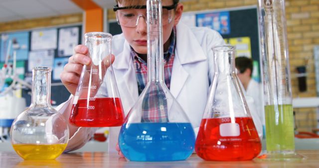 Young boy wearing safety goggles conducting a chemistry experiment with colorful beakers in a laboratory setting. Useful for educational content, science promotions, children's educational material, or science-related advertisements.
