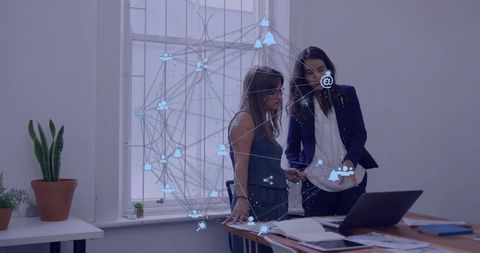Women collaborating on laptop with holographic network overlay, pointing at documents
