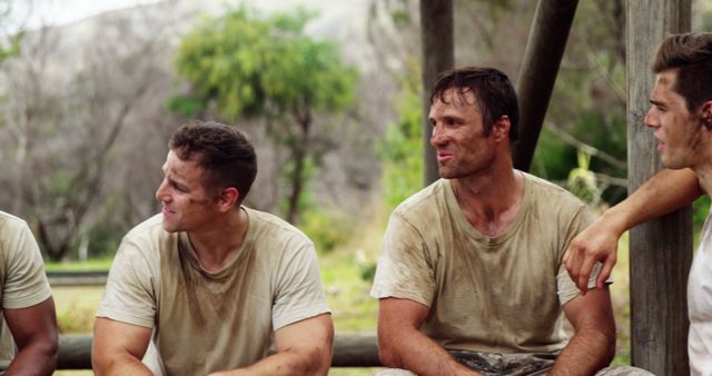Caucasian men sit together outdoors, with copy space. They appear relaxed and muddy, suggesting a break from an outdoor activity.