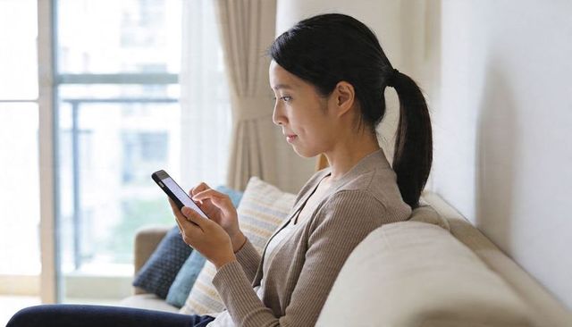 Asian woman scrolling smartphone on sofa in bright modern apartment