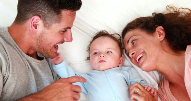 Parents lying in bed with their baby, smiling and engaging in playful interaction. Perfect for concepts of family bonding, nurturing environment, joy of parenthood, or promoting family-related products.