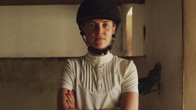 Caucasian woman dressed in equestrian gear wearing a riding hat, standing with arms crossed in stable, displaying confident demeanor and subtle smile. Could be used in advertisements for equestrian sports, horse riding lessons, or representing confident individuals in sports. Ideal for promotional material, sports and lifestyle articles.