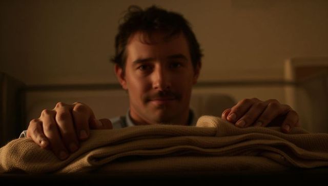 Young man gripping folded beige blanket on metal bed rail in dim bedroom, warm lamplight