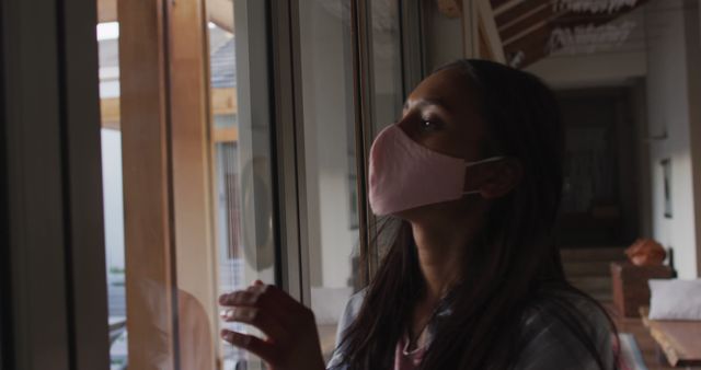 Young girl wearing pink face mask looking out a window indoors. She appears thoughtful while peering outside, suggesting themes of quarantine, safety measures during the COVID-19 pandemic, or the feeling of isolation. Useful for articles, blogs, or awareness campaigns about health precautions, mental health during isolation, or COVID-19 related content.