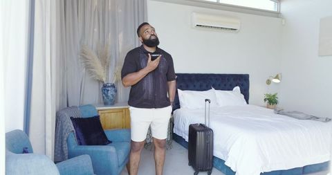 African American Man Relaxing in Modern Bedroom with Smartphone and Suitcase