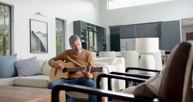 Mature man sitting on a comfortable sofa, playing acoustic guitar and enjoying leisure time in a modern, bright living room with elegant furnishings. Ideal for use in articles about hobbies, relaxation at home, music practice, and enjoying life.