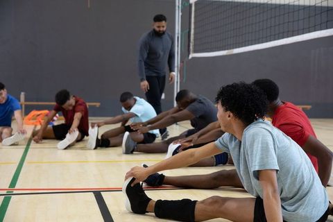 Youth male volleyball team stretching and warming up on indoor gym court with coach guidance