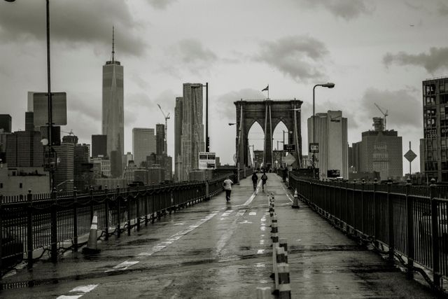 The scene captures pedestrians walking across a wet Brooklyn Bridge on a rainy day, with the New York City skyline visible in the background. The overcast weather and wet pavement add a moody and atmospheric tone to the urban landscape. Ideal for use in articles or websites about travel, urban exploration, architecture, and the dynamic spirit of New York City.