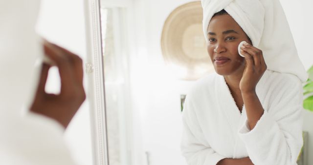 Image of happy african american woman in robe looking at mirror and cleansing face. beauty treatment and skin care routine concept.