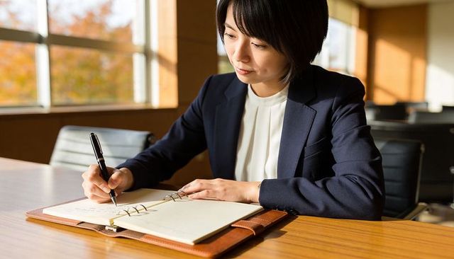 Businesswoman writing in leather ring notebook at boardroom table in warm daylight
