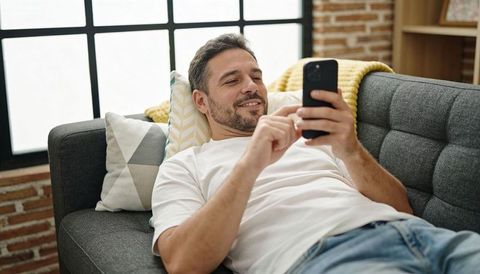 Man relaxing on tufted gray sofa scrolling smartphone in cozy industrial living room
