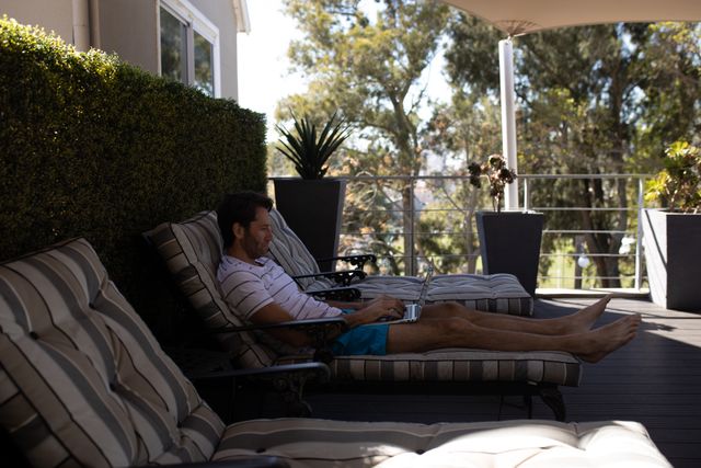 Caucasian man enjoying leisure time on a hotel terrace, lying on a sunlounger with a laptop. Ideal for themes related to relaxation, summer vacations, remote work, technology use in leisure, and outdoor activities.