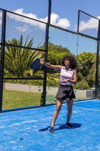 Playing padel on bright blue court wearing smartwatch and black skort holding paddle