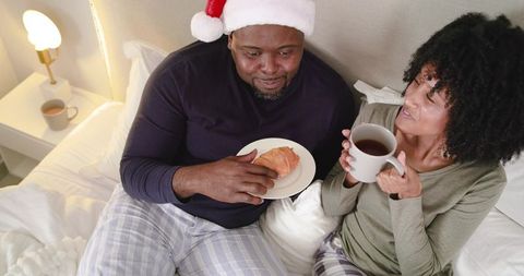 African american couple sharing cozy christmas morning breakfast in bed wearing santa hat