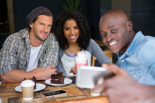 Happy young man with friends taking selfie at wooden table in coffee shop