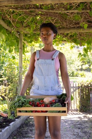 Teenage african woman holding crate of vegetables under pergola wearing denim overalls