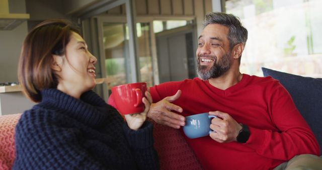 Happy diverse couple sitting in living room drinking coffee and talking. Spending quality time at home.