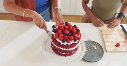Diverse friends cutting three-layer red velvet cake with fresh berries at kitchen counter