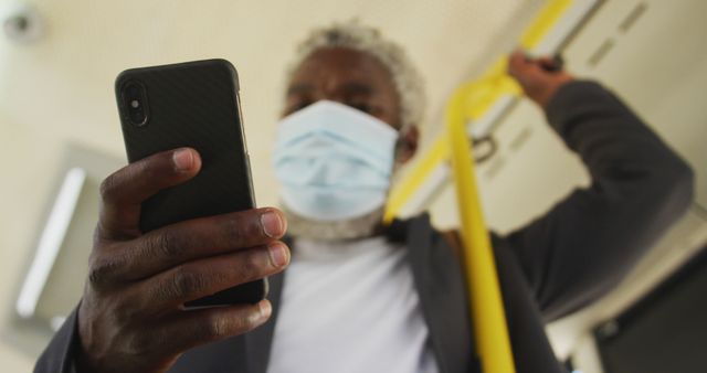 Close-up view of a senior man wearing a face mask, holding onto a pole in public transportation, and using his smartphone. Depicts safety measures during travel, everyday technology use, and commuting. Ideal for illustrating public health campaigns, technology adaptation among elderly, or commuting lifestyle.