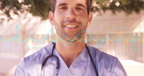 Smiling doctor wearing blue scrubs and stethoscope standing under tree canopy