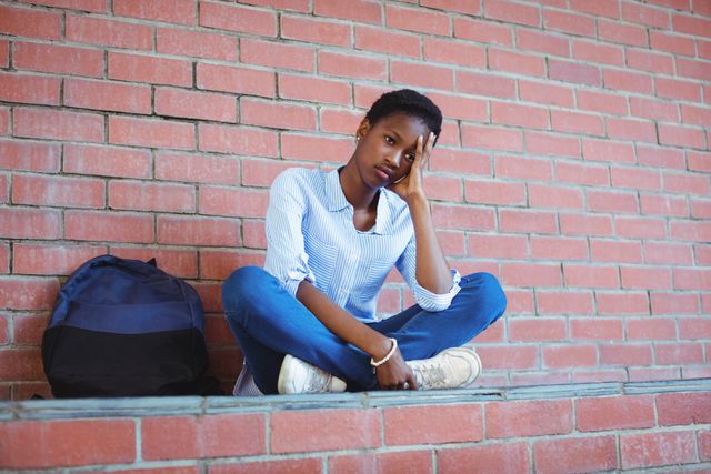 Sad teenage girl sitting alone against a brick wall at school campus, looking stressed and thoughtful. Surrounding includes a backpack suggesting a school setting. Ideal for illustrating themes of education, stress, mental health, teenage emotions, and solitude among students.